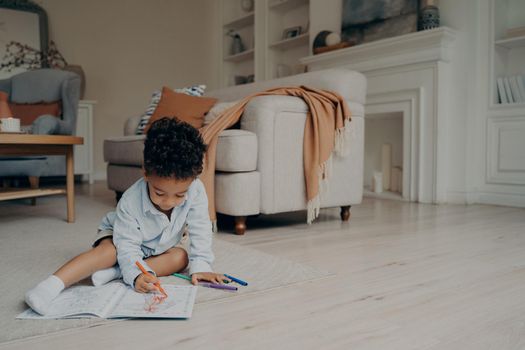 Little mixed race kid boy drawing in coloring book while spending leisure time alone at home