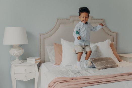 Energetic afro american kid jumping with lollipop on bed mattress