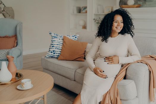 Pregnant afro american woman looking at window while sitting on sofa at home