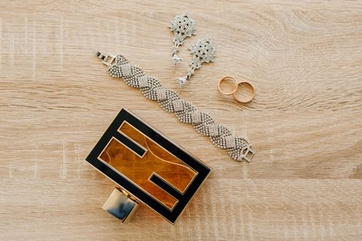 Perfume bottle, bracelet, stud earrings and wedding rings on a light wooden surface.