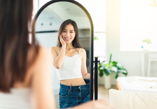 A young woman wearing large jeans before losing weight, looking at the figure in front of the mirror, is very happy that she has lost weight successfully, showing a very confident smile