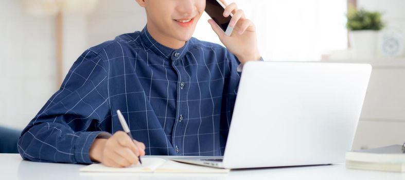 Young asian man talking phone and work from home with laptop computer, freelance male writing on notebook and speak on smartphone, stay home, domestic life, business and communication concept.