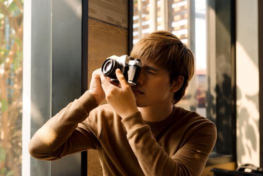 Male amateur photographer in eyeglasses focusing to make photos out of window sitting on windowsill.Young bearded man resting in coworking space and taking pictures for portfolio on vintage camera