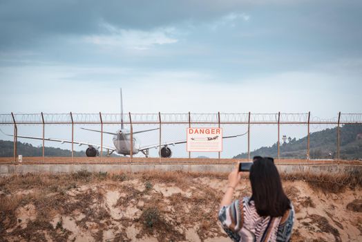 Tourist Woman is Capturing Photos The Airplane While Take off Landing on Runway Track From Outside The Airport. Asian Tourist Woman Having Fun With Photographic Vehicle Aircraft Beside Airport Fence.