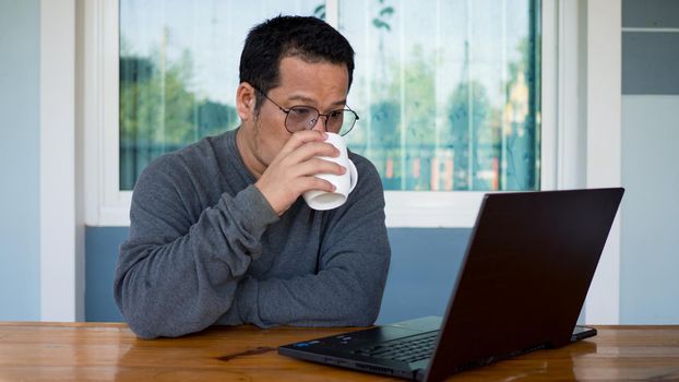 Asian men wear long sleeves and eyeglasses.He is drink a coffee and Sitting at home working on a computer on a wooden table. Work from home concept.