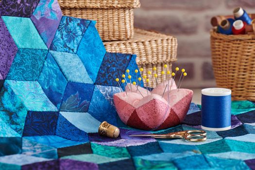 Pincushion, spool of thread, scissors, thimble and fragment of tumbling blocks quilt on baskets background