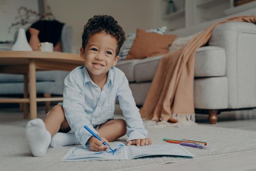 Little afro kid boy sitting on floor and drawing with colorful felt tip pens
