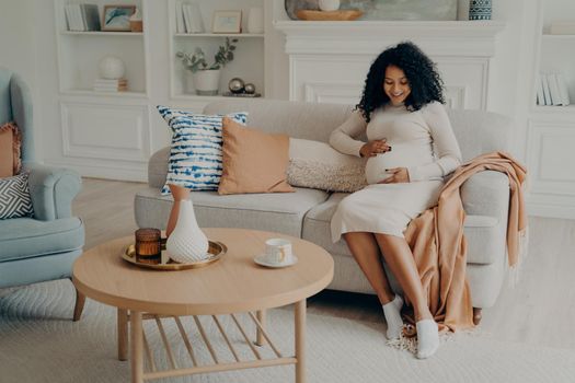 Pregnant afro american woman sitting on couch and spending time at home