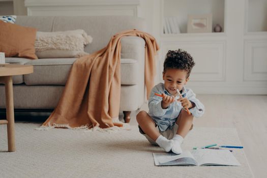 Cute afro american boy kid with colouring book sitting on floor in cozy living room