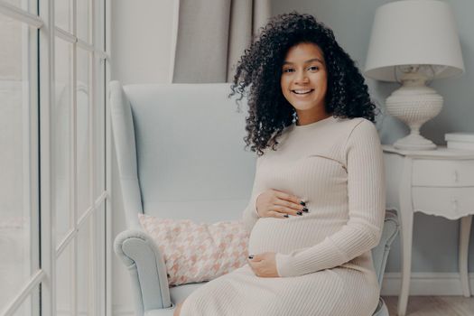 Afro American adorable smiling pregnant lady in beige dress sits in white armchair