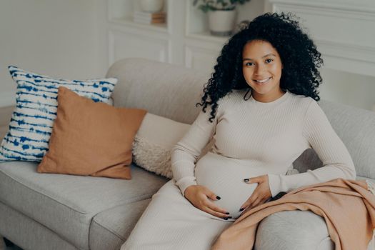 Lovely Afro American future mother sitting on sofa and enjoying happy pregnancy time