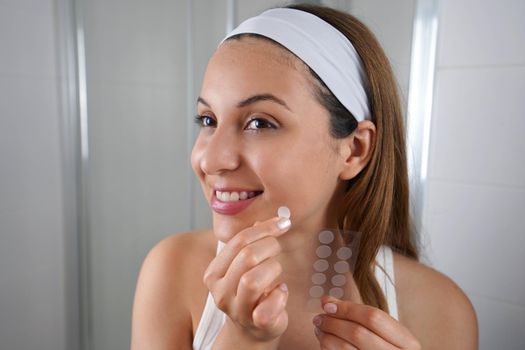 Beautiful smiling girl applying acne treatment patch on a pimple in bathroom