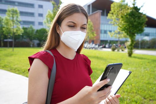Busy female manager with FFP2 KN95 protective mask checking her smartphone when walking outside her enterprise office