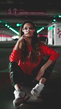 Pretty young woman sitting on car parking. Wearing stylish urban outfit