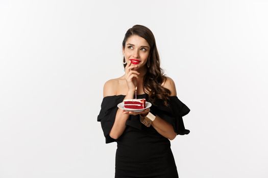 Celebration and party concept. Dreamy woman in black dress making wish, thinking and holding birthday cake with candle, standing over white background